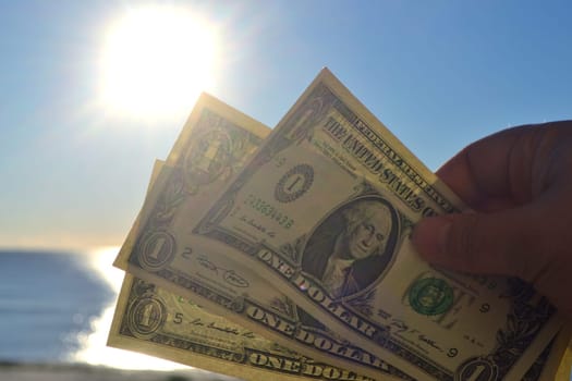 Person holding in hand paper bills of dollars with face value of one dollar on background of brightly shining sun, blue sky and calm blue sea. One-dollar paper banknotes on backdrop of blue sea sky