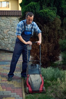 Full length portrait of a professional Caucasian male gardener trimming grass lawn using electric cordless mower in the courtyard. Landscaping Industry Theme. House and garden maintenance concept