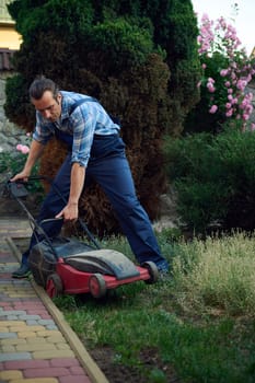 Low angle view of Caucasian man gardener in blue work uniform, working at garden. Professional landscaper using electric lawn mower, cutting grass with modern gardening equipment. Landscaping Industry