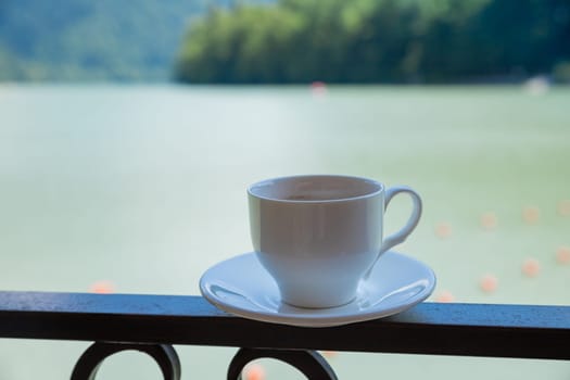 Cup of hot coffee on terrace with beautiful view of Alpine mountain landscape at background with small lake downhill in valley.