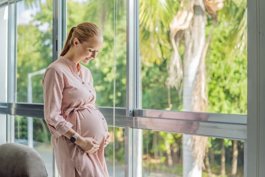 Portrait of young pregnant woman standing near window holding hands on her belly at modern home with copy space. Pregnancy, motherhood, expectation and tenderness concept.