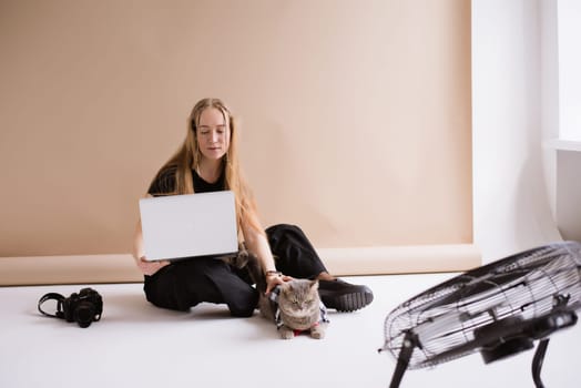 A business woman freelancer in a black is working, typing on a laptop apple and sitting on a white floor. blonde assistant in video production with computer MacBook and camera with cat