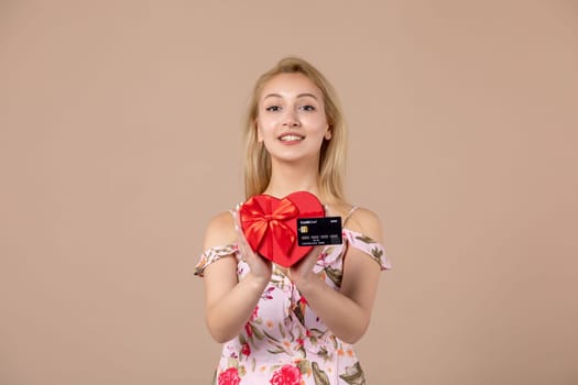 front view young female posing with red heart shaped present and bank card on brown background feminine money march equality woman sensual horizontal