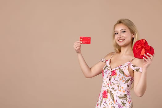 front view young female posing with red heart shaped present and bank card on brown background feminine money march horizontal equality sensual