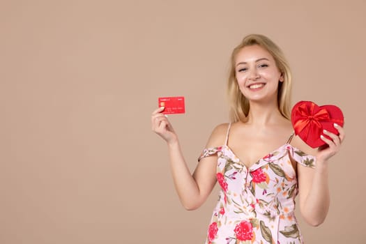 front view young female posing with red heart shaped present and bank card on brown background feminine money march horizontal equality woman