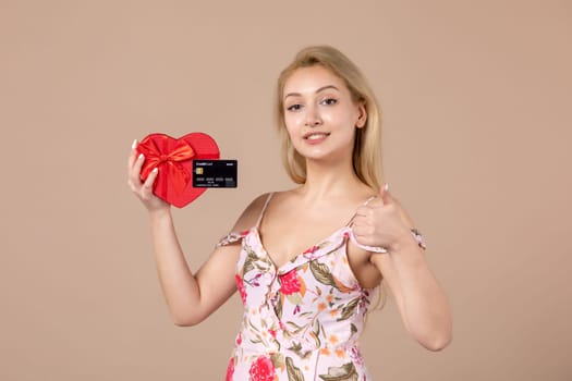 front view young female posing with red heart shaped present and bank card on brown background feminine money march sensual horizontal equality
