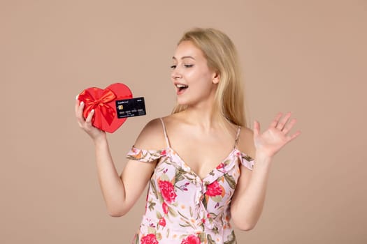 front view young female posing with red heart shaped present and bank card on brown background feminine money march woman sensual horizontal