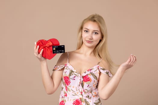 front view young female posing with red heart shaped present and bank card on brown background feminine money march woman sensual horizontal equality