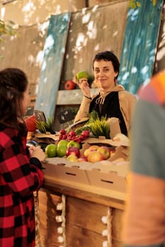 Smiling local vendor selling organic fresh apples at farmers market stand, wearing apron and talking to customers. Young woman holding bio natural locally grown farming products.