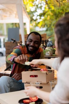 Happy young African man customer tasting organic fruits and vegetables at farmers market, smiling black guy sitting at table with local vendor discovering apple varieties while visiting food fair