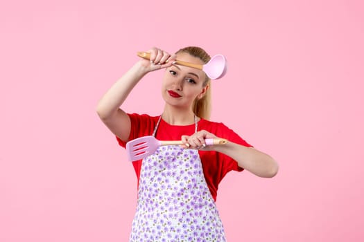front view young pretty housewife in cape with spoons on pink background uniform profession cooking worker job wife duty occupation