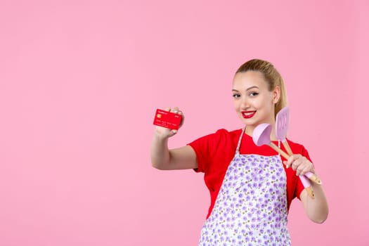 front view young housewife in cape holding spoons and bank card on pink background duty money worker horizontal wife uniform job cutlery