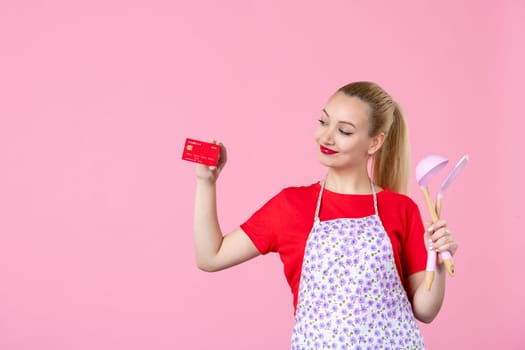 front view young housewife in cape holding spoons and bank card on pink background occupation cutlery horizontal job wife duty profession money uniform