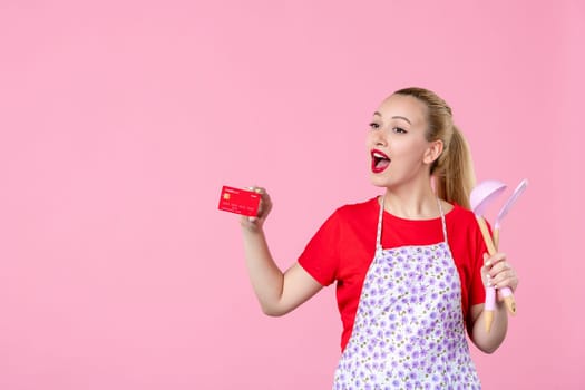 front view young housewife in cape holding spoons and bank card on pink background occupation duty money worker horizontal wife profession job cutlery