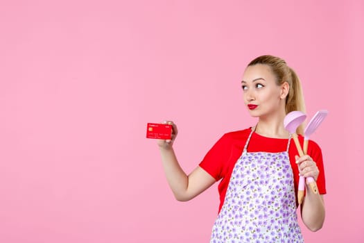 front view young housewife in cape holding spoons and bank card on pink background occupation duty worker cutlery horizontal wife profession money job