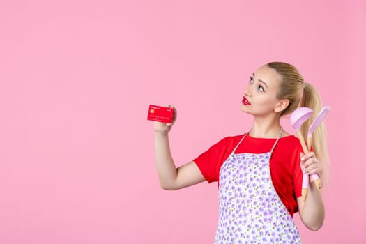 front view young housewife in cape holding spoons and bank card on pink background occupation duty worker cutlery horizontal wife profession money uniform job