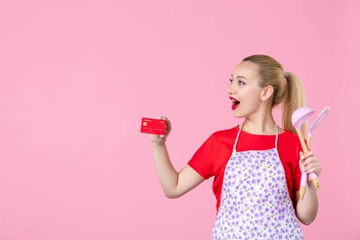 front view young housewife in cape holding spoons and bank card on pink background occupation duty worker cutlery horizontal wife profession uniform job