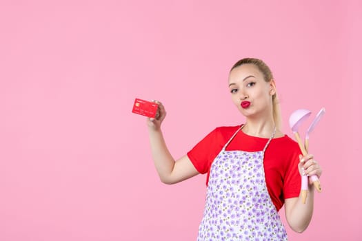 front view young housewife in cape holding spoons and bank card on pink background occupation worker horizontal job wife duty profession money uniform