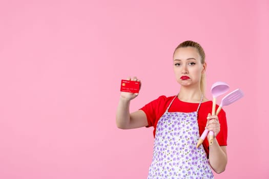 front view young housewife in cape holding spoons and bank card on pink background profession duty money horizontal wife uniform job cutlery worker