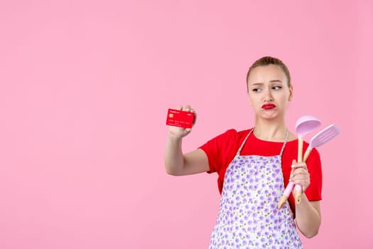 front view young housewife in cape holding spoons and bank card on pink background profession occupation duty horizontal wife uniform job cutlery worker