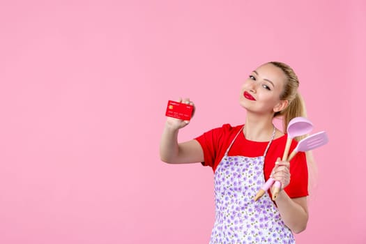 front view young housewife in cape holding spoons and bank card on pink background profession occupation duty money horizontal wife job cutlery worker