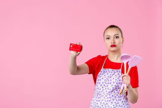 front view young housewife in cape holding spoons and bank card on pink background profession occupation duty money wife uniform job cutlery horizontal