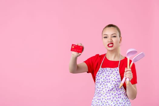 front view young housewife in cape holding spoons and bank card on pink background profession occupation duty money wife uniform job cutlery worker