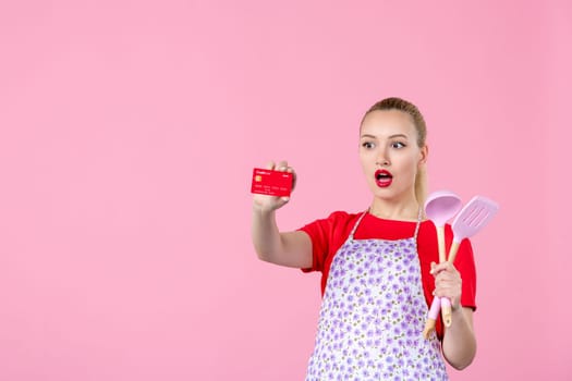 front view young housewife in cape holding spoons and bank card on pink background profession occupation duty money wife uniform job cutlery worker