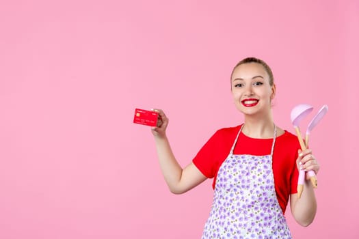 front view young housewife in cape holding spoons and bank card on pink background worker cutlery uniform horizontal occupation job duty profession