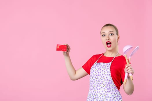 front view young housewife in cape holding spoons and bank card on pink background worker cutlery uniform horizontal occupation job wife duty profession