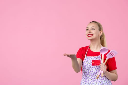 front view young housewife in cape holding spoons and red bank card on pink background occupation profession job horizontal wife money duty workers uniform