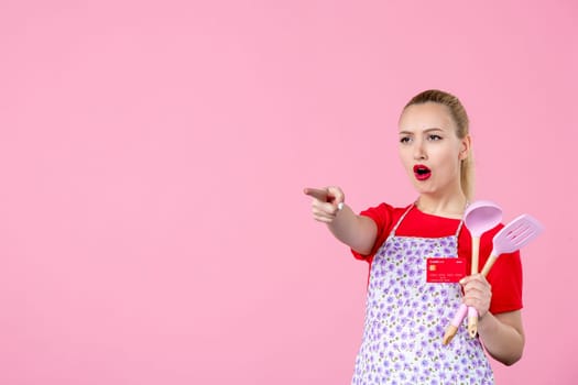 front view young housewife in cape holding spoons and red bank card on pink background occupation profession job horizontal wife money worker uniform