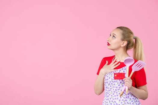 front view young housewife in cape holding spoons and red bank card on pink background occupation uniform profession job horizontal wife duty worker