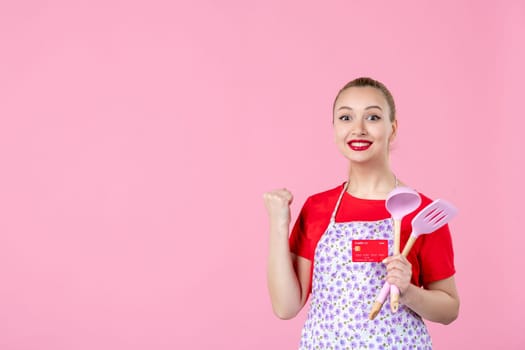 front view young housewife in cape holding spoons and red bank card on pink background occupation uniform profession job horizontal wife money