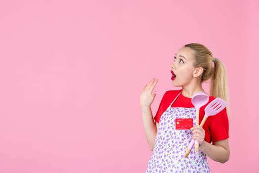 front view young housewife in cape holding spoons and red bank card on pink background occupation uniform profession job horizontal wife money duty workers