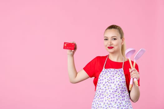 front view young housewife in cape holding spoons and red bank card on pink background profession money occupation duty uniform horizontal wife