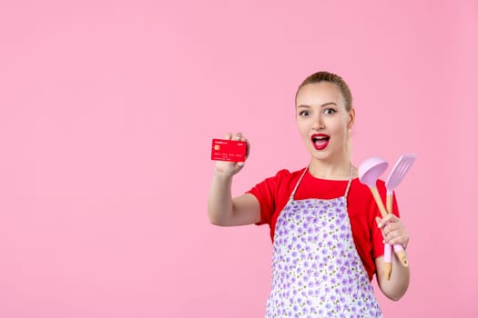 front view young housewife in cape holding spoons and red bank card on pink background profession money occupation duty uniform job cutlery horizontal wife