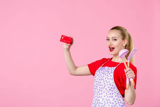 front view young housewife in cape holding spoons and red bank card on pink background profession money occupation duty uniform job cutlery worker horizontal