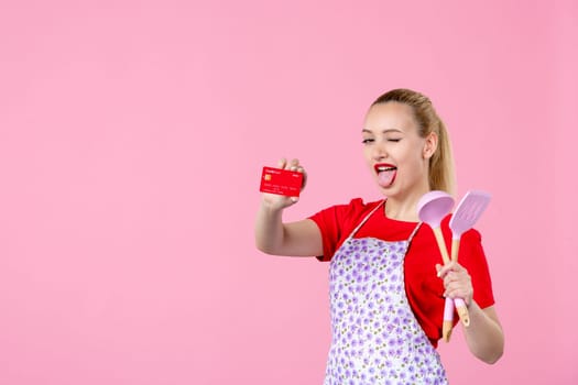 front view young housewife in cape holding spoons and red bank card on pink background profession occupation duty money uniform job cutlery horizontal wife