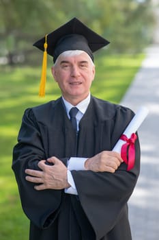 Portrait of an elderly man in a graduation gown and with a diploma in his hands outdoors. Vertical