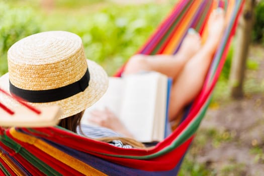 Young woman reading book while lying in comfortable hammock at green garden.