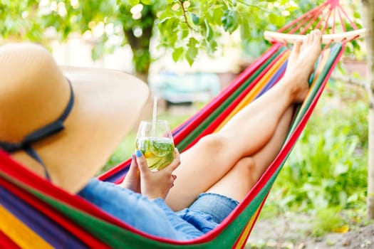 Young woman drinking a cocktail while lying in comfortable hammock at green garden.