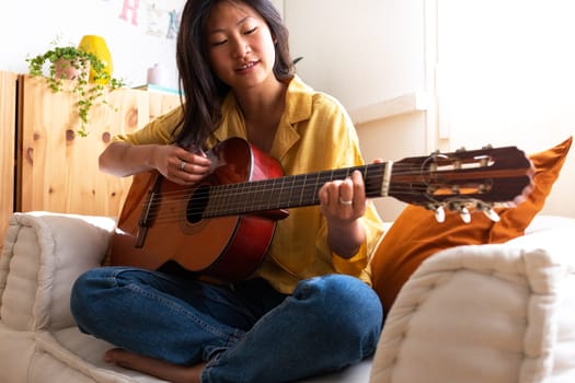Happy teen asian girl composing song sitting on couch. Practicing chords. Playing acoustic guitar. Female singer songwriter. Musician and hobbies concept.