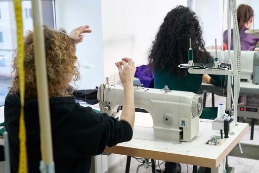 Young dressmaker woman sews clothes on sewing machine. Smiling seamstress and her hand close up in workshop. Focus on sewing machine and tissue.