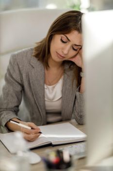 A pensive young woman sitting alone and worriedly write something down in a notebook while using a computer.