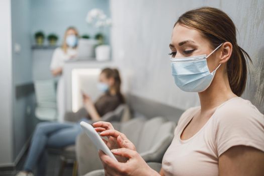 An attractive young woman wearing face mask and using her cellphone while sitting on a chair in waiting room at dentist's office.