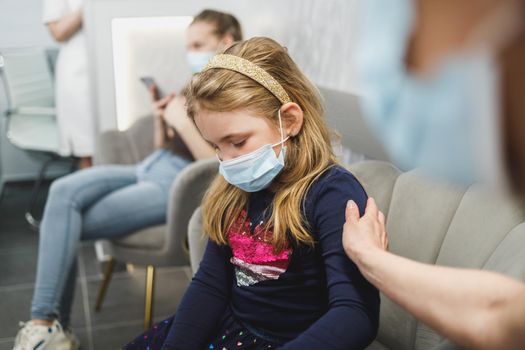 Sad little girl and her mom with face mask in waiting room at dentist's office.