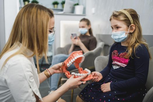 Female dentist and little girl wearing protective face masks while holding model of teeth and communicating in waiting room.