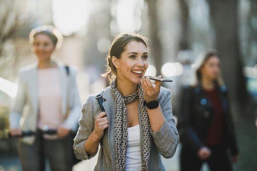 A smiling young business woman talking voice message on a smart phone on her way to work.