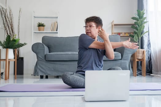 Active mature woman doing stretching exercise in living room at home. Fit lady stretching arms and back while sitting on yoga mat.